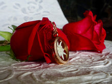 2 roses are laying on top of white handmade paper. Leaning against the closer rose if a locket terrarium pendant with a white edelweiss inside. 