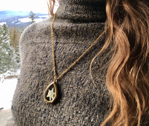 a young woman with long brown hair wears a gold locket that is made of glass and is a teardrop shape. The locket is see-through. Inside the locket is a white edelweiss flower that has been preserved. In the background is snow, trees and mountains. The edelweiss locket is handmade by Pressed Wishes. 