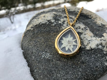 A gold teardrop locket is see-through and is made of glass. Inside the locket is a white edelweiss that has been preserved. The locket is on a gold chain and is laid on a rock surrounded by snow. The edelweiss locket is handmade by Pressed Wishes. 