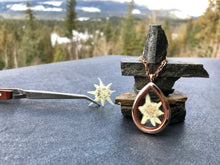 A white edelweiss flower is placed inside a rose gold and glass see-through locket necklace. The locket necklace stands against a mini inukshuk. In the background is forest, snow and mountains. The locket is handmade by Pressed Wishes, a canadian floral artist. 