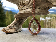 A white pressed edelweiss is inside a glass and rose gold see-through locket in a teardrop shape. The locket is set against a piece of driftwood. The locket is handmade by Pressed Wishes. 