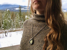 A young woman with long golden hear wears a wooly turtleneck and a silver locket. The locket is a teardrop shape and has a single white edelweiss inside. The edelweiss is in the glass terrarium locket. The background of the picture contains snow and, trees and mountains. Edelweiss locket is handmade by Pressed Wishes. 