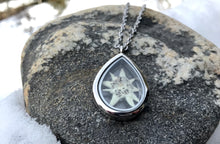 a white preserved edelweiss is in a silver stainless steel and glass locket that is a teardrop shape. The locket lays on a grey rock in a patch of snow. The item is handmade by Pressed Wishes 
