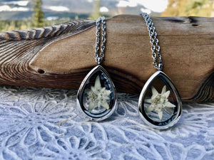Two silver and glass lockets lay against a piece of drift wood. Inside the lockets are a single white edelweiss flower that has been pressed and placed inside. The locket is on a long chain that drapes of the wood.  The jewellery is handmade by Pressed Wishes