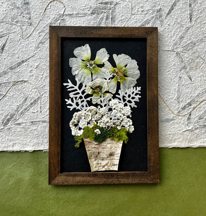 A Bouquet of pressed flowers including Hollyhock, Pearly Everlasting, and moss are contained in a birch planter pot and framed with a wooden frame. The pressed botanical artwork is by Pressed Wishes. 