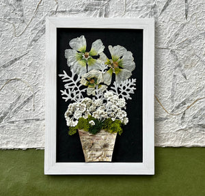 A Bouquet of pressed flowers including Hollyhock, Pearly Everlasting, and moss are contained in a birch planter pot and framed with a white wooden frame. The pressed botanical artwork is by Pressed Wishes.