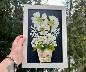 A hand holds a handmade picture containing a bouquet of pressed flowers including Hollyhock, Pearly Everlasting, and moss are contained in a birch planter pot and framed with a wooden frame. The pressed botanical artwork is by Pressed Wishes.