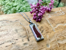 a pink Veronica flower is encased in a resin and presented in a vertical bar pendant. The pendant lays on a piece of wood. 