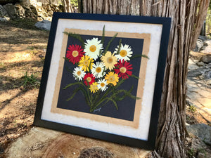 A bouquet of colourful pressed Shasta daisies are arranged on layers of f handmade paper and framed with a solid wood handcrafted frame. This real pressed daisy picture is made by botanical artist, Pressed Wishes, from Mabel Lake, BC, Canada. The pressed botanical picture leans against the trunk of a tree. 